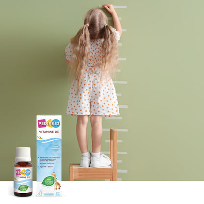 PEDIAKID Vitamina D3 picături - fetiță care își măsoară înălțimea lângă perete, alături de flaconul cu picături